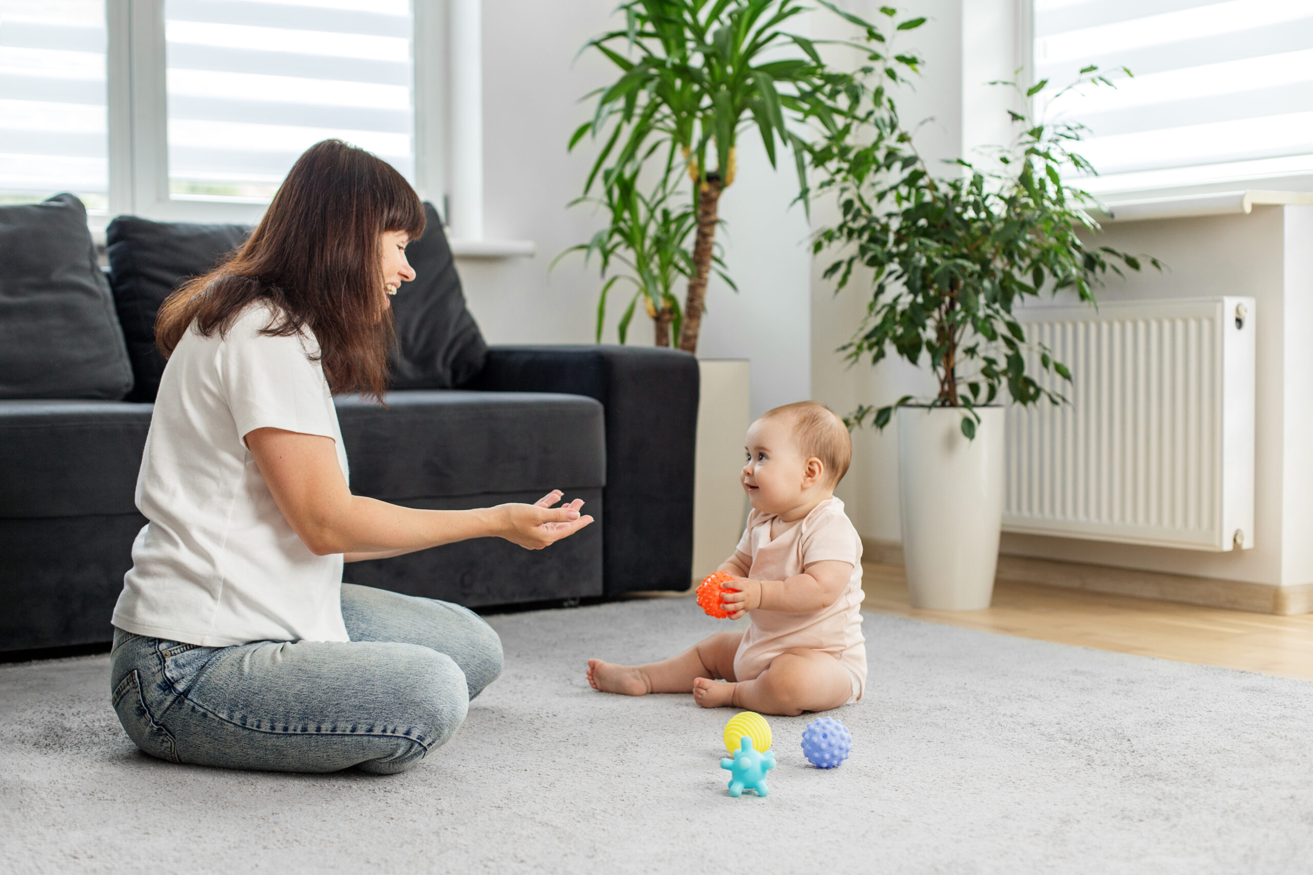 Smiling baby holds sensory ball while sitting on living room carpet, as mother interacts warmly, showing family love and playful bonding.