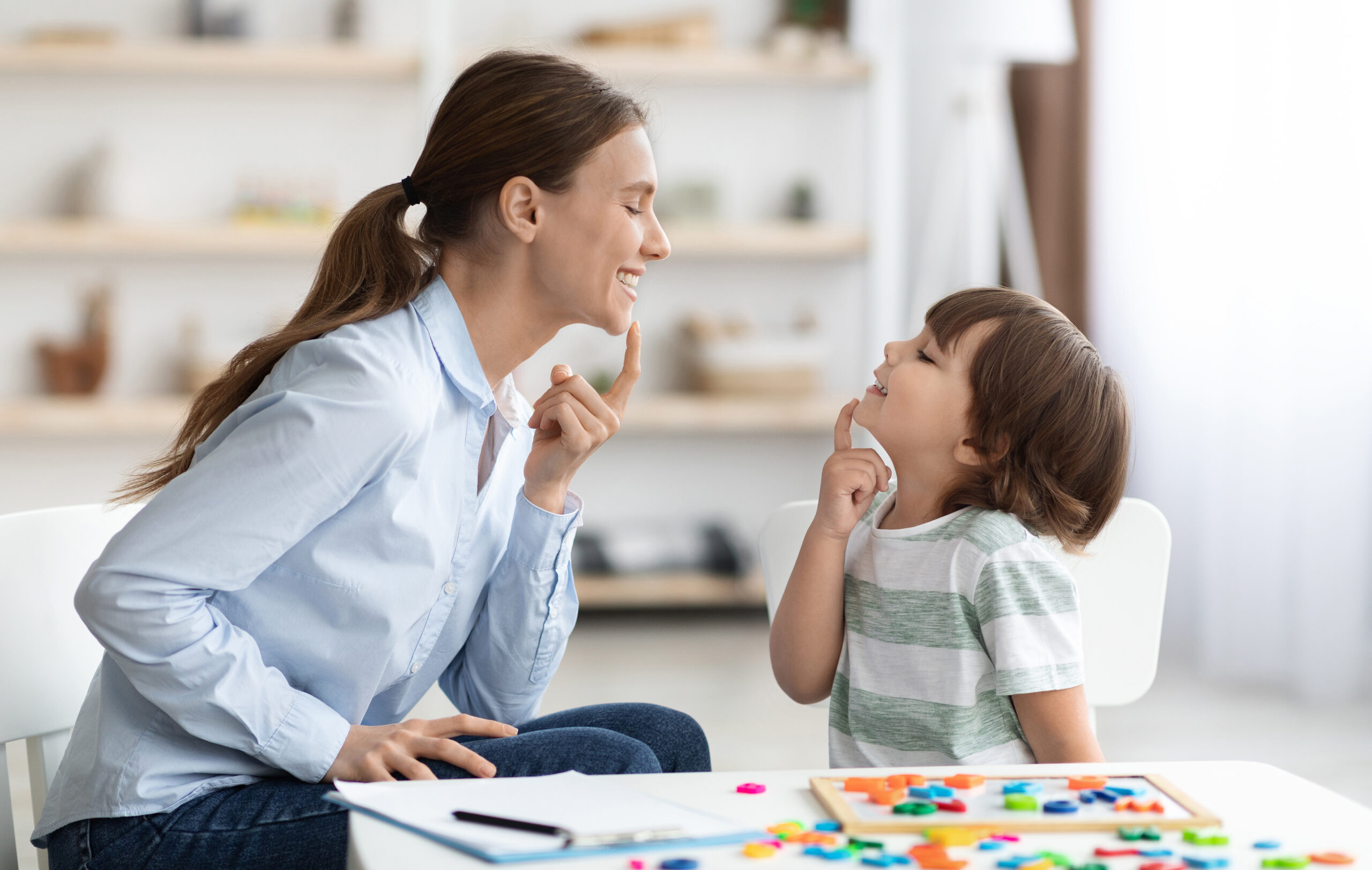 Speech training for kids. Professional woman specialist training with little boy at cabinet, teaching him right articulation exercises, side view